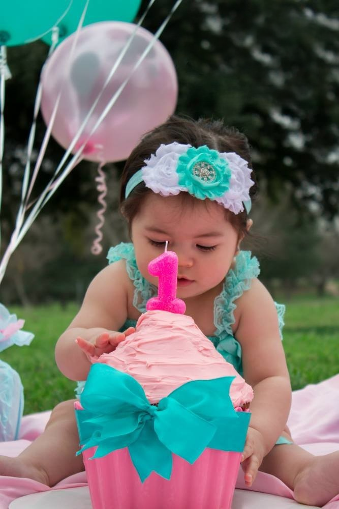 A one year old baby girl touches pink frosting on a giant cupcake while sitting near balloons.