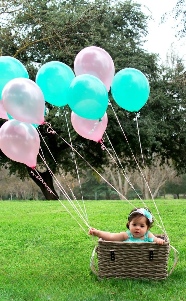 A one year old baby girl sits in a basket and holds onto balloons while taking a photo.