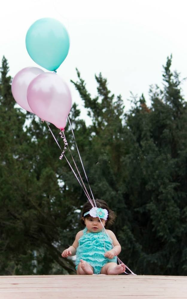 A one year old baby girl holding balloons, poses for a photo.