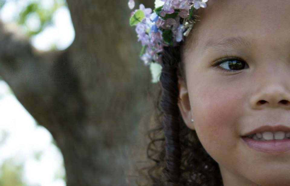 A young girl smiles for the camera while standing by a tree, with half of her face fully in view.