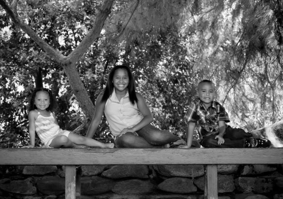 Three children sit on a bench and pose in a park.