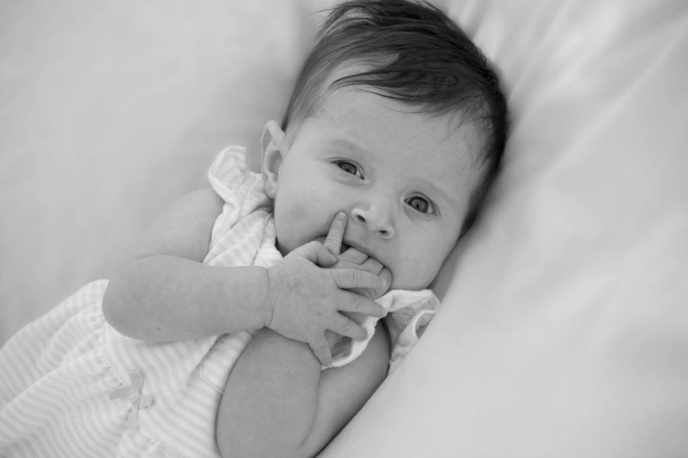 An infant girl sucks on its fingers and gazes into the camera lens.