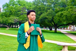 Graduate in green cap and gown holds diploma while standing on a tree-lined campus walkway.