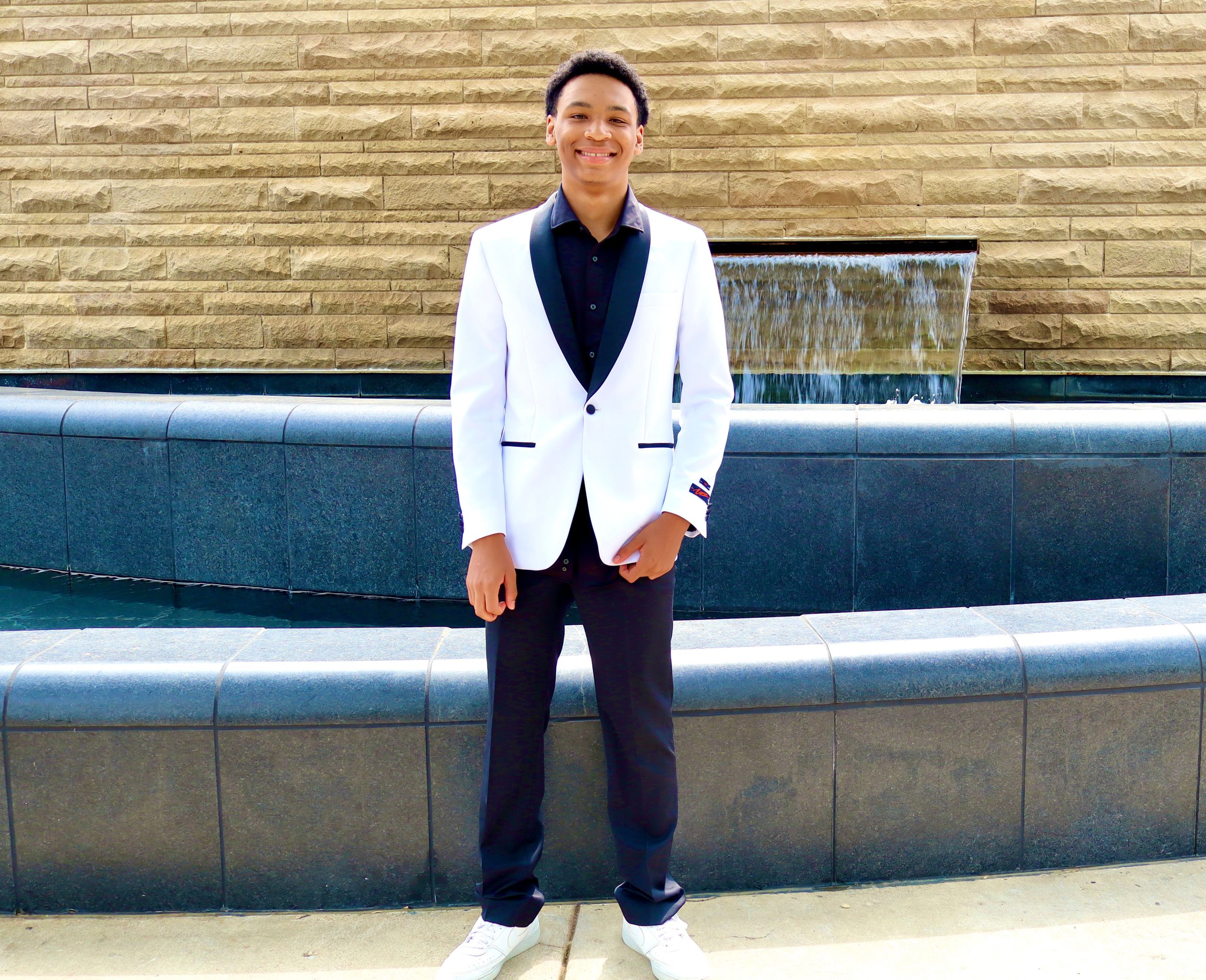 Young man in white tuxedo jacket and dark pants stands confidently near a fountain outdoors.