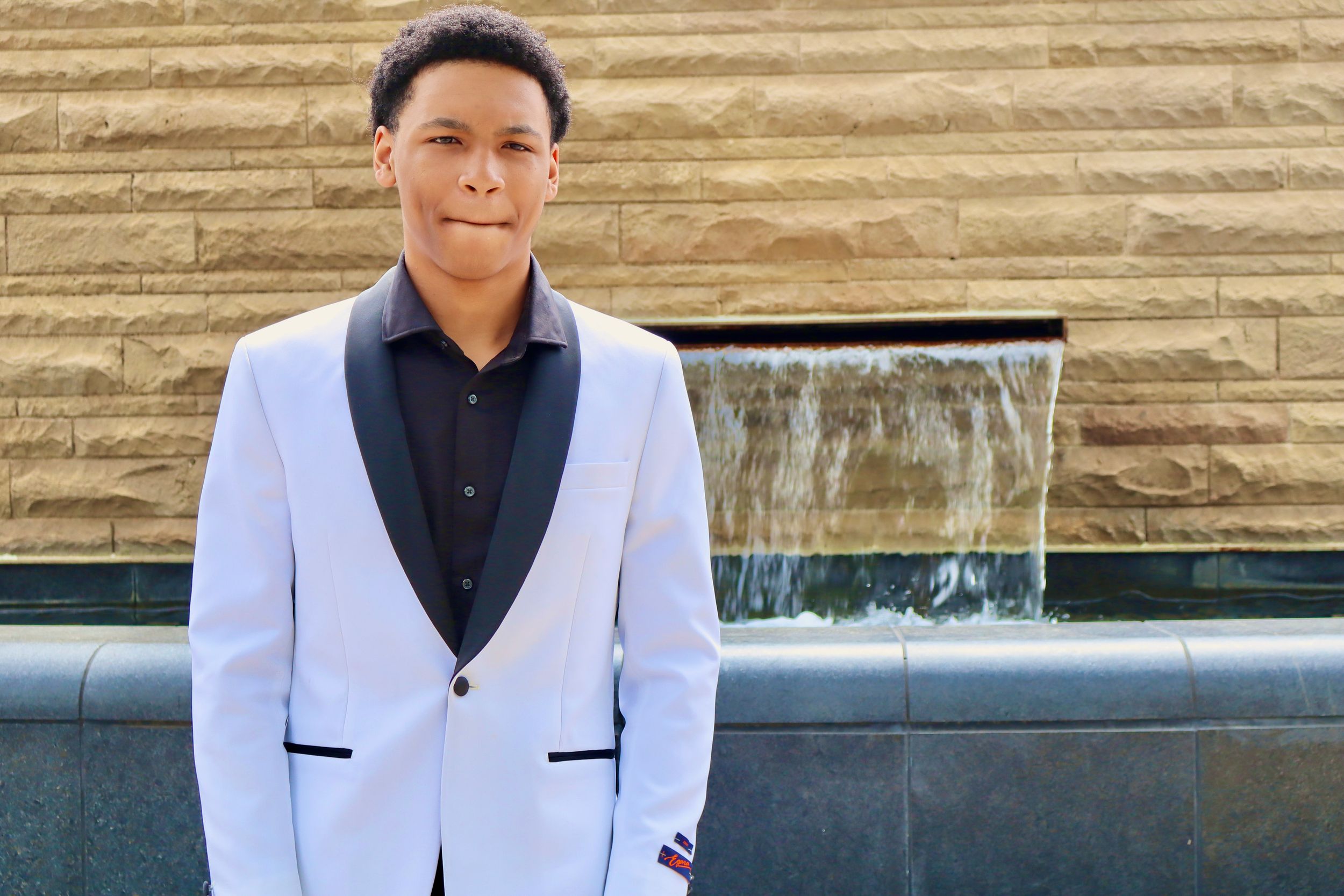 Young man in white tuxedo jacket poses in front of a stone wall with a waterfall feature.
