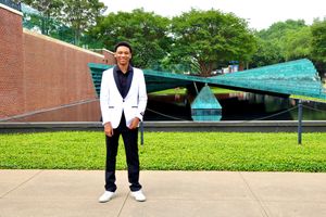 Young man in white blazer stands smiling outdoors near modern architectural water feature.