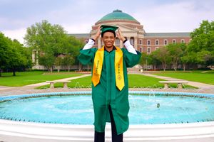 Graduate in green cap and gown celebrates in front of a university fountain on a cloudy day.