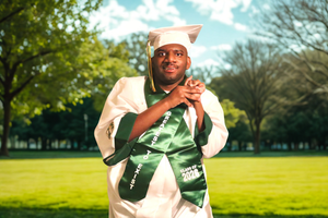 Graduate in white cap and gown with green stole poses outdoors on green lawn with trees.