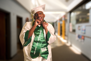 Smiling graduate in white cap and gown with green stole poses in sunny school hallway.