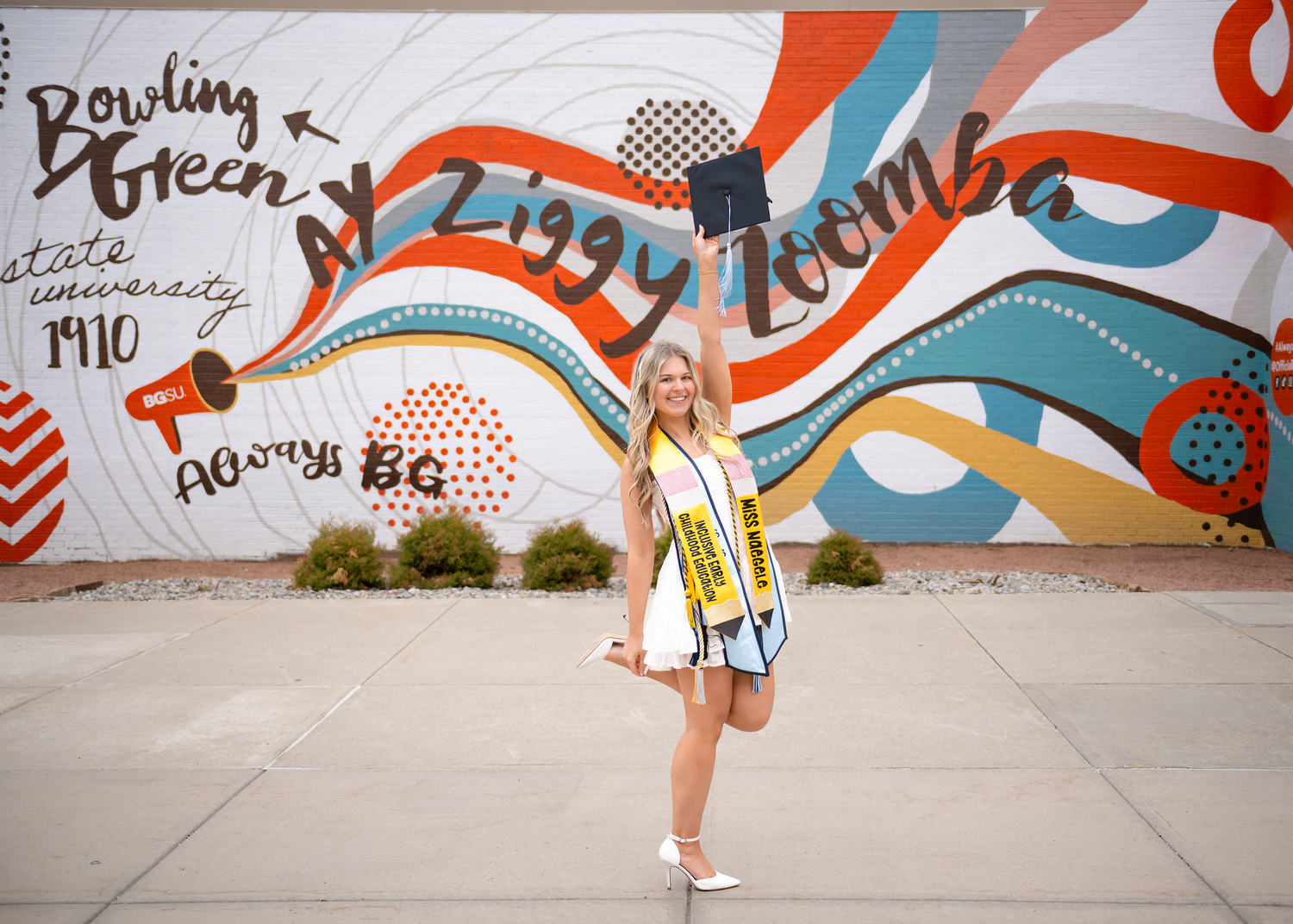 Young woman in white dress with sash poses before colorful Bowling Green State University Ziggy Zoomba mural.