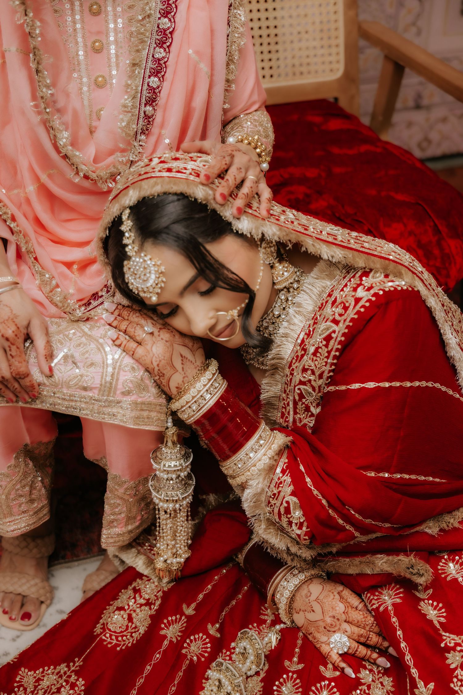 A bride in red lehenga with gold embroidery rests her head on an elder's lap during an Indian wedding ceremony.