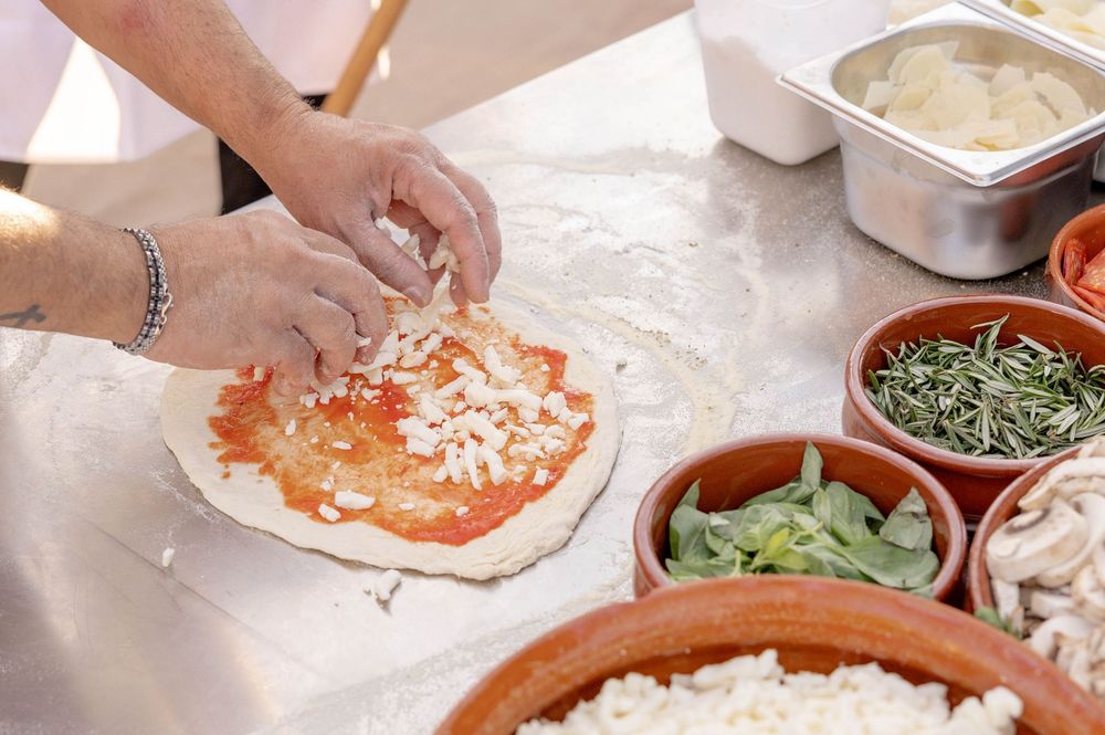 Chef hands spreading cheese on a fresh pizza dough with tomato sauce, surrounded by herb bowls on a flour-dusted surface.