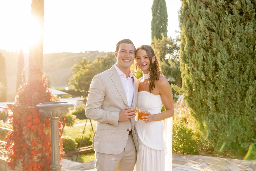 Couple smiling outdoors at golden hour, man in tan suit, woman in white dress, holding drinks with cypress trees behind.