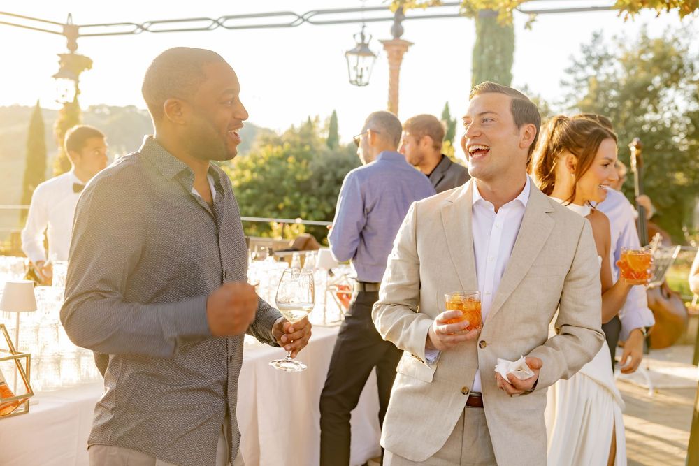Two men laughing and holding cocktails at an outdoor wedding reception in warm golden sunlight.