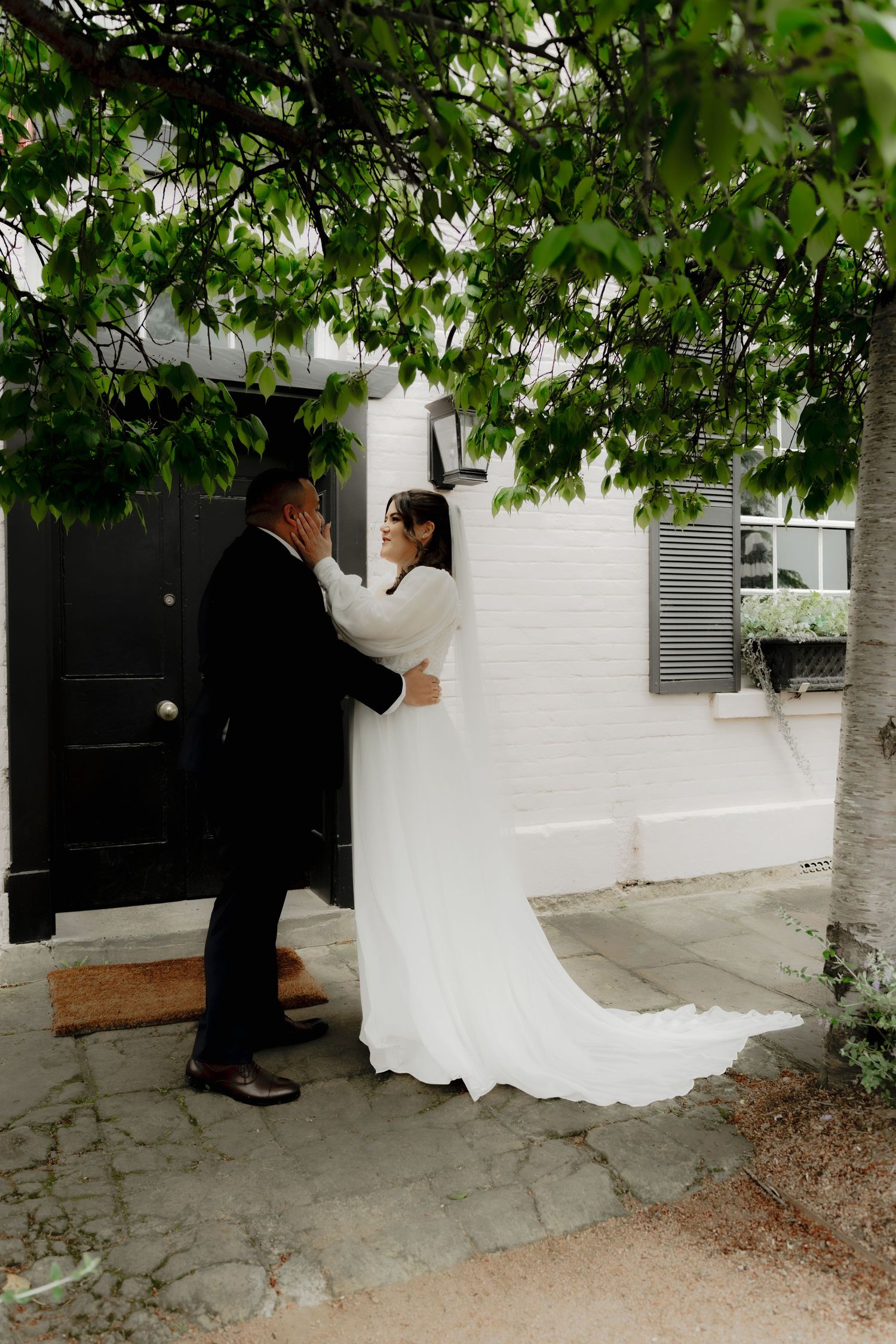 Bride in white gown with train and groom in black suit share a tender moment at a black door.