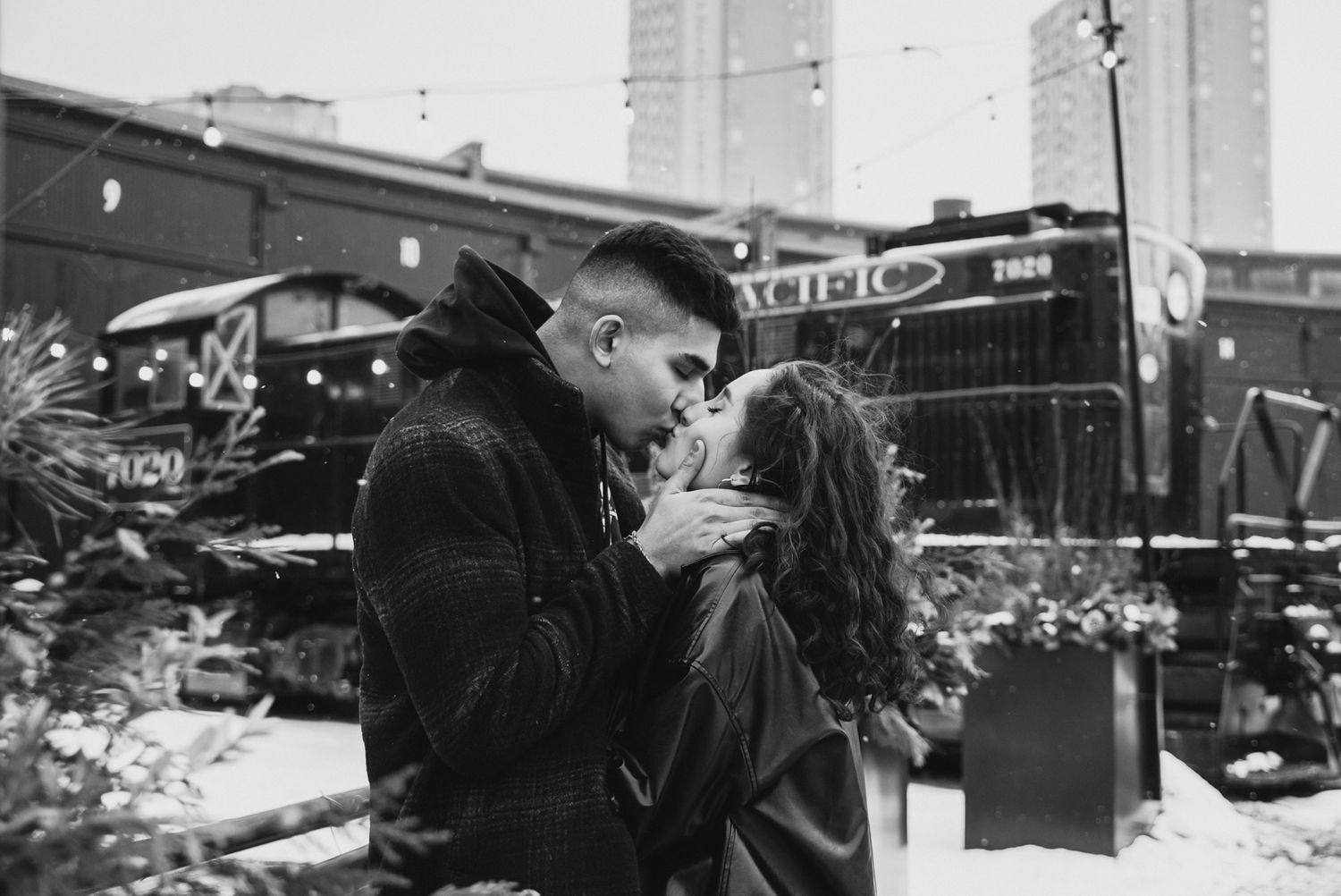 Couple sharing a romantic kiss in the snow outside a vintage train in a city setting, black and white photo.