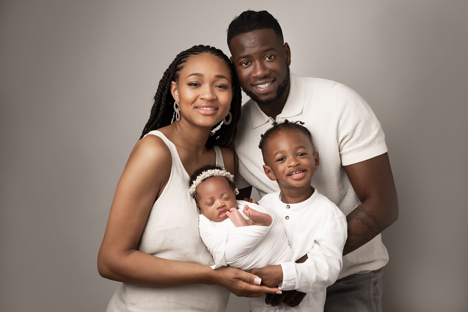 A joyful family of four poses for a studio portrait, parents holding a newborn and young child, all dressed in white.