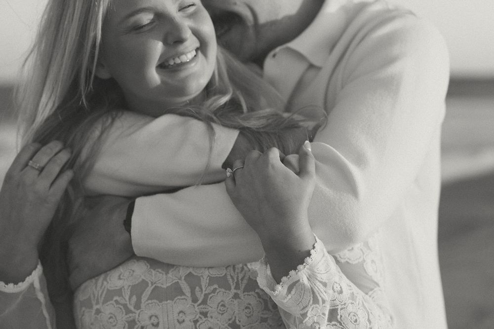 Black and white photo of a smiling woman being embraced from behind by a man, holding hands romantically outdoors.