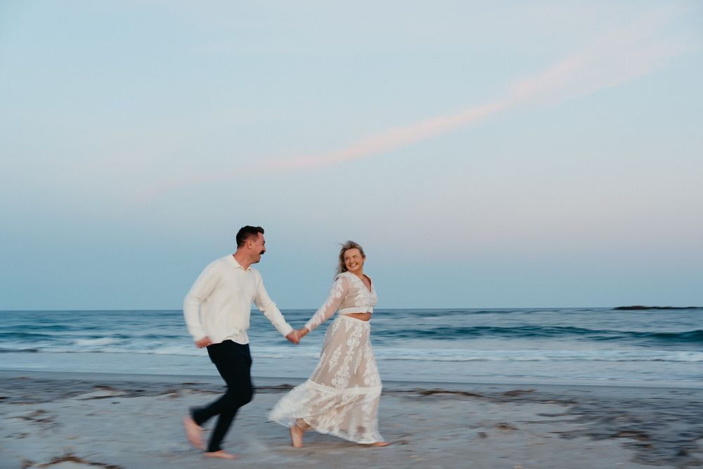 Couple holding hands running on a sandy beach at sunset, woman in lace dress, man in white sweater. Wrightsville Beach Couples Photography
