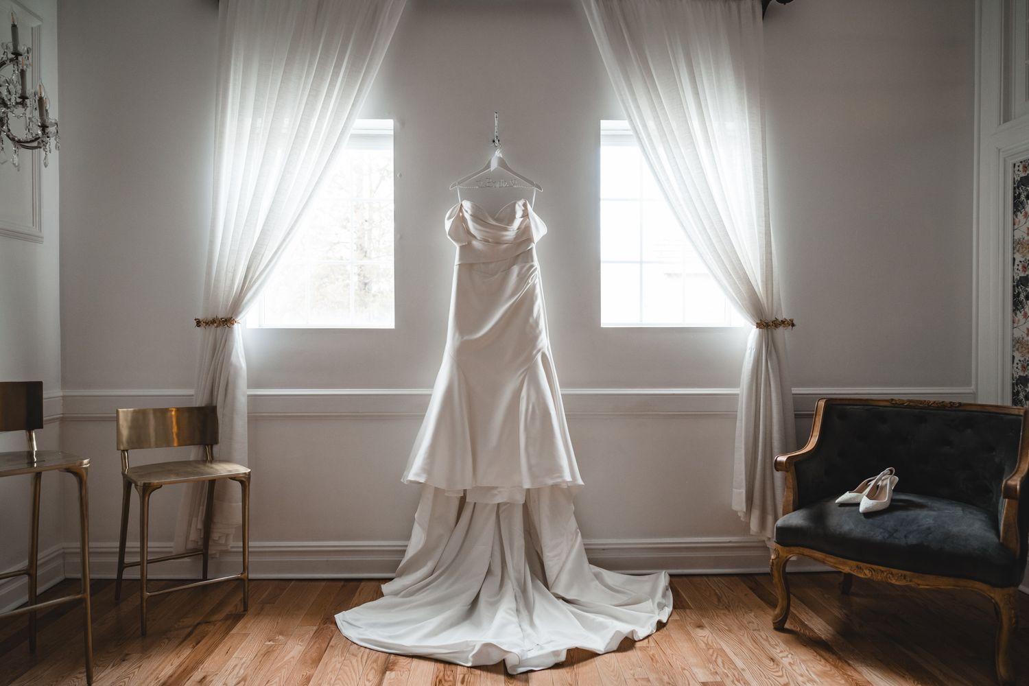 Elegant white wedding dress hanging between two windows in a classic room with hardwood floors.