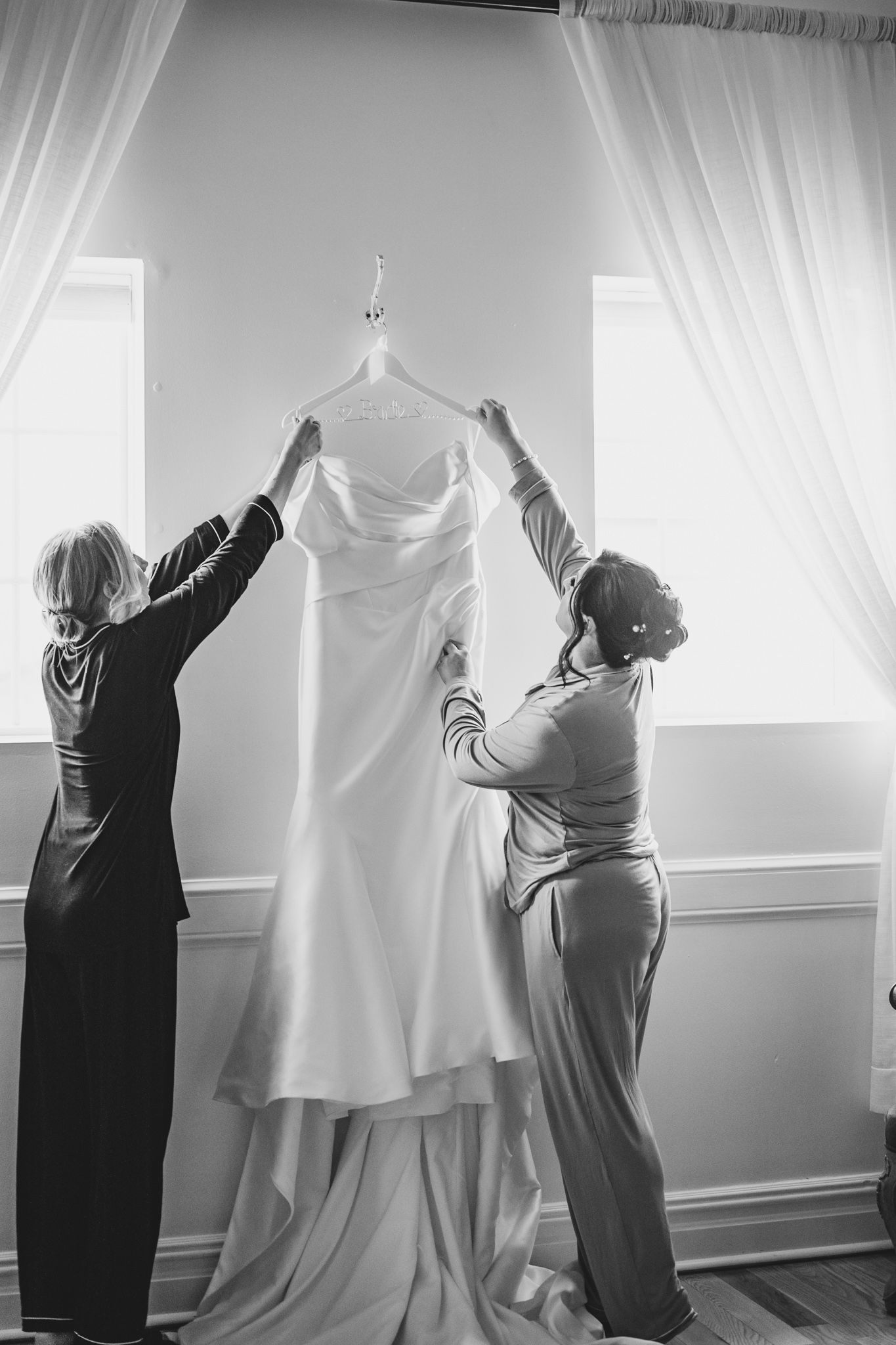 Two women lift a white wedding gown on a hanger in a bright room with sheer curtains.