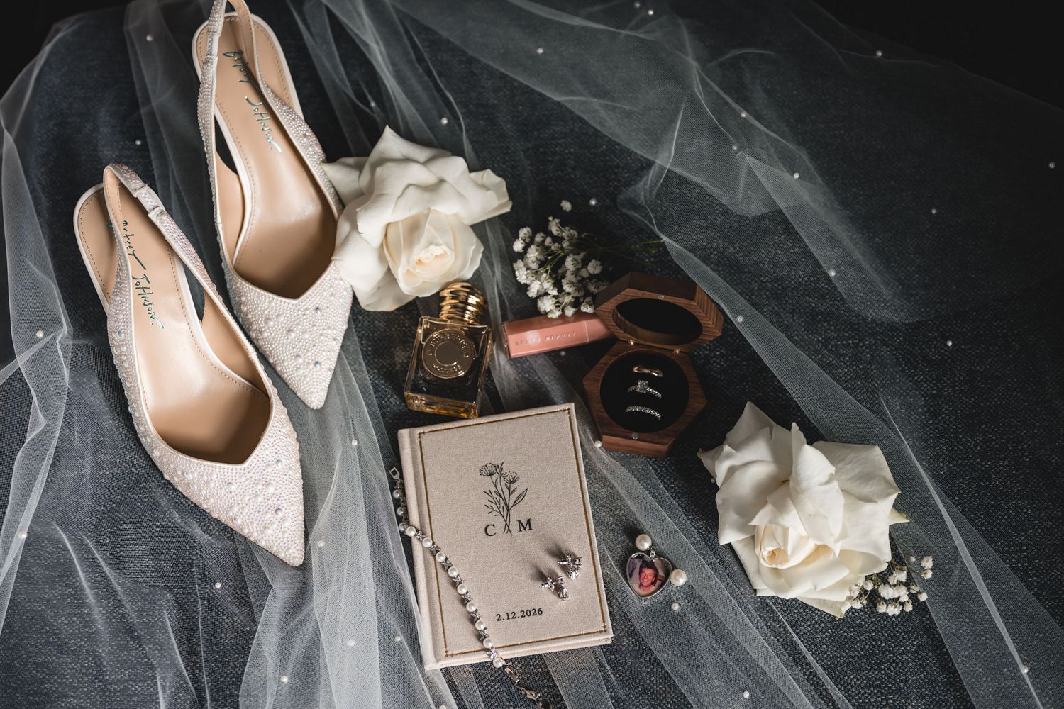 Elegant wedding flat lay with lace heels, white roses, perfume, ring box, and monogrammed card on sheer veil.