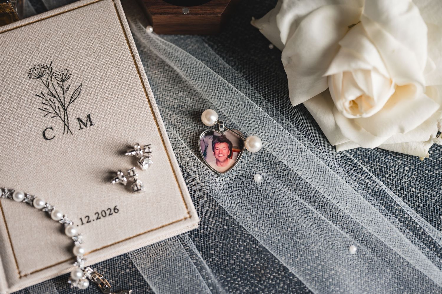Wedding flat lay with monogrammed box, pearl jewelry, photo charm, and white rose on a veil for a 2026 bride.