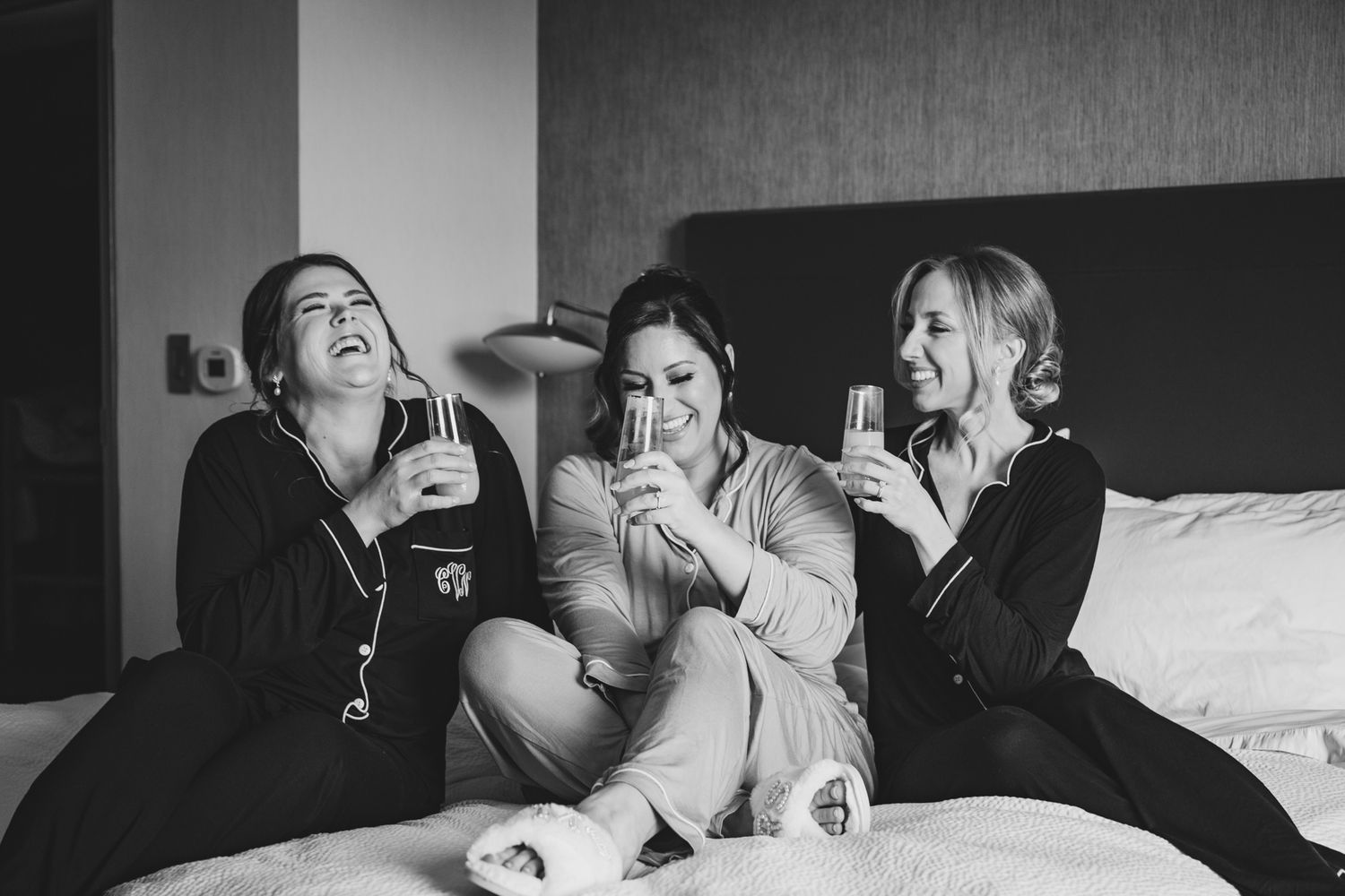 Three bridesmaids laughing and drinking champagne together on a hotel bed in black and white photo.