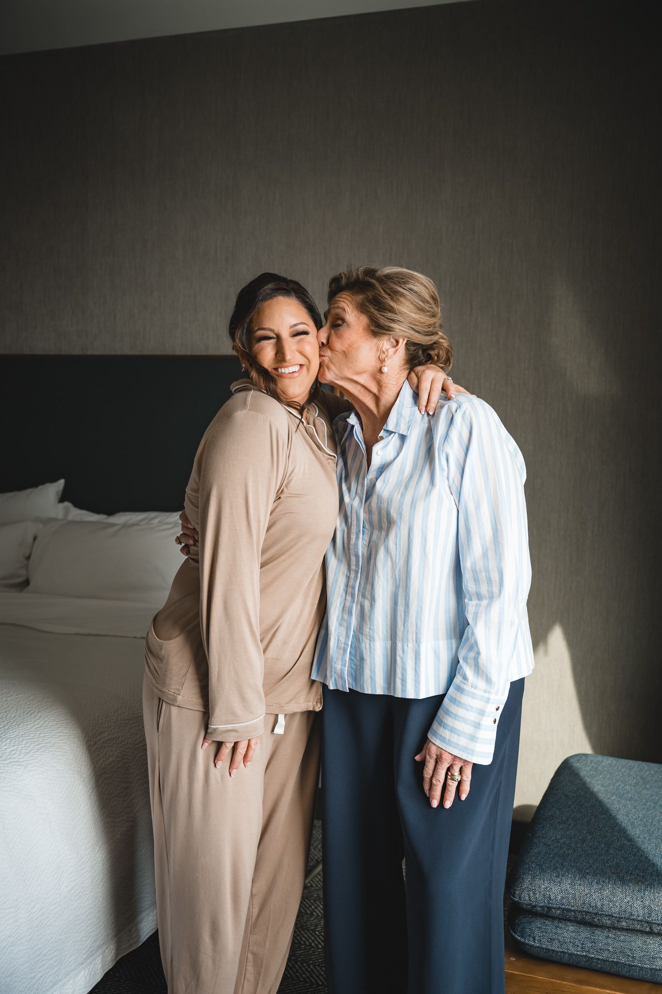 Two women sharing a tender moment as an older woman kisses a younger smiling woman on the cheek in a hotel room.