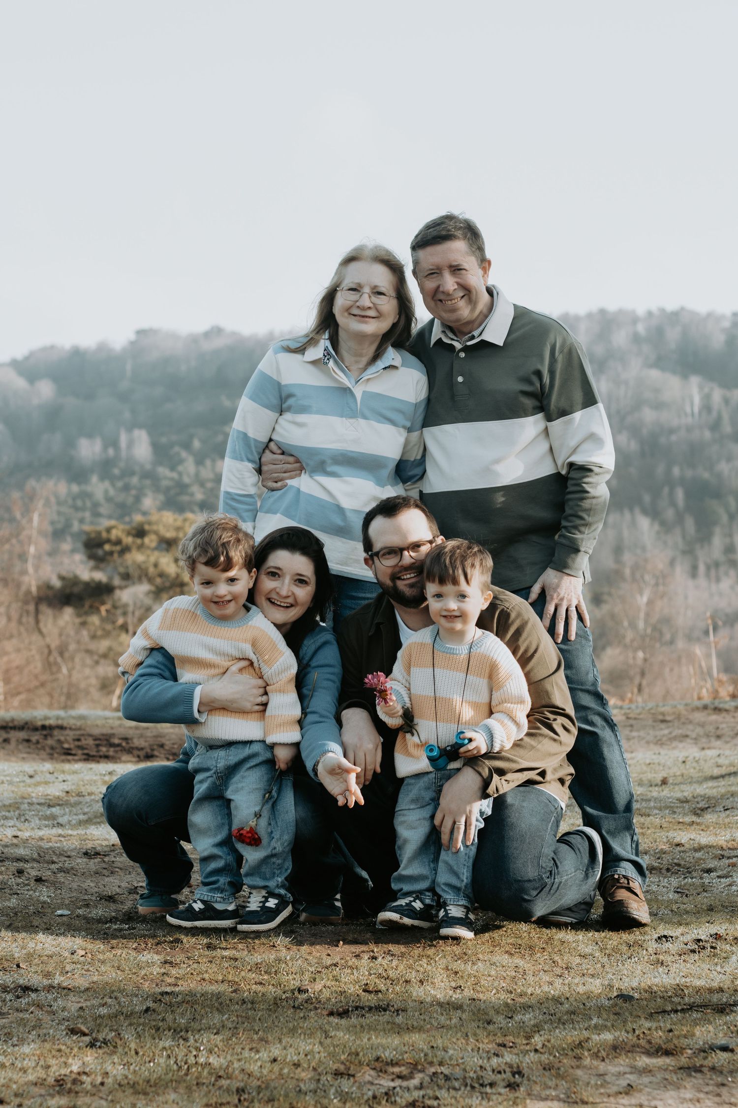 Multi-generational family of six poses outdoors for a portrait, smiling together against a natural hillside backdrop.