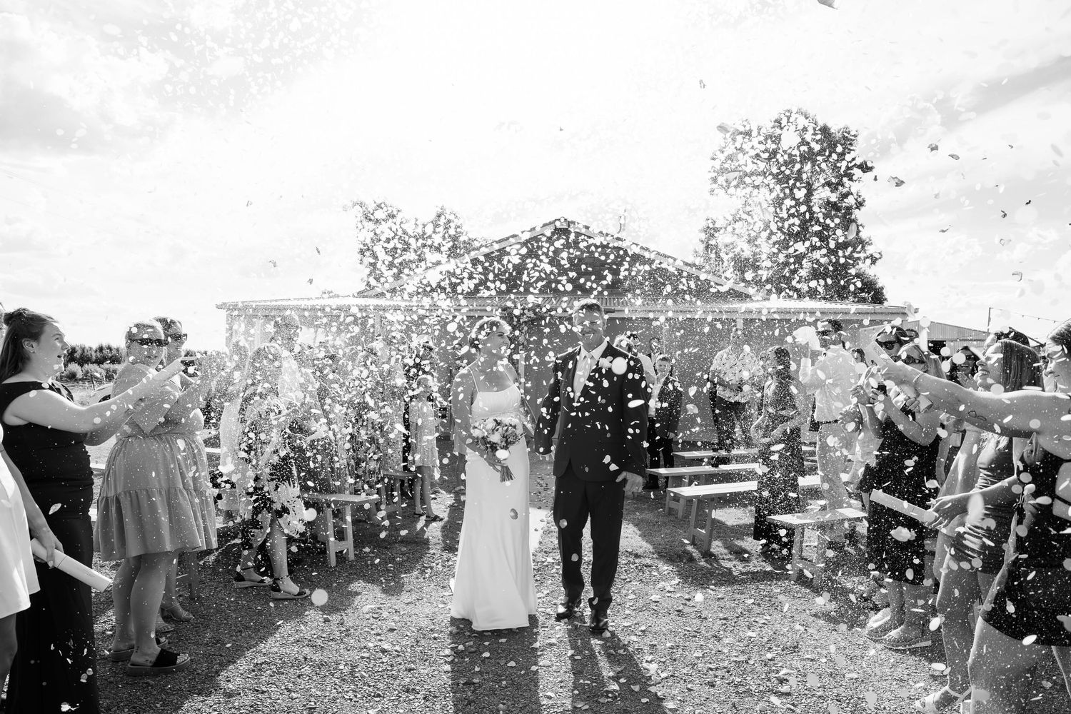 Black and white photo of bride and groom walking through confetti shower after outdoor wedding ceremony.
