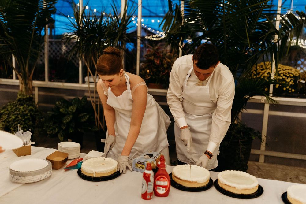 Two people in white aprons cutting wedding cakes at a table in a greenhouse venue at night.