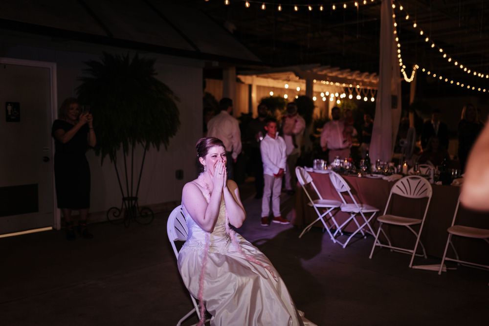Emotional wedding guest in gold gown covers mouth while seated at outdoor evening reception with string lights.