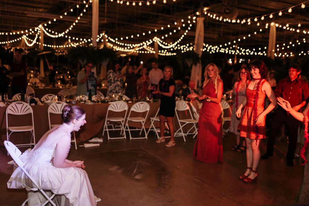 Bride seated at wedding reception with guests dancing under string lights in decorated barn venue.