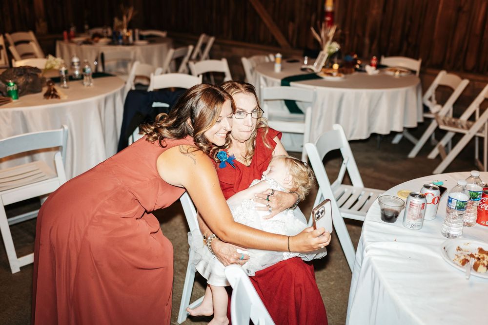Two women in red dresses embrace a child at a wedding reception with white chairs and decorated tables.