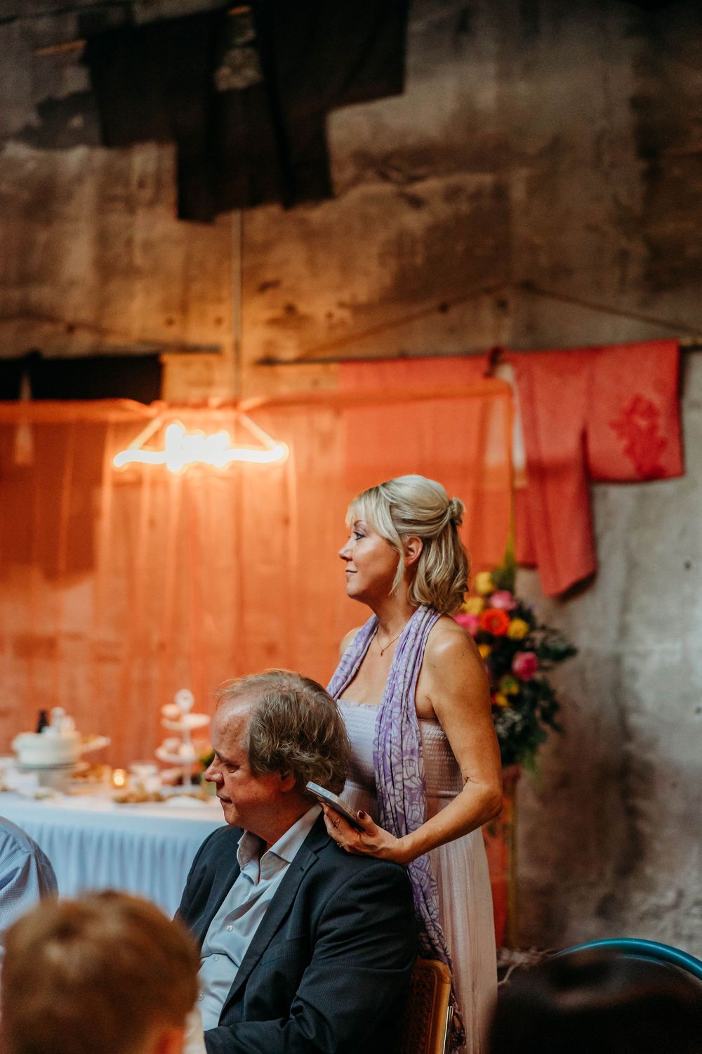 Elegant wedding guest in lavender dress standing behind seated man at rustic barn reception.