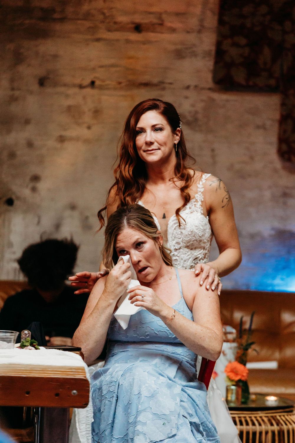 Bride in lace gown comforts emotional woman in blue dress wiping tears at wedding reception.