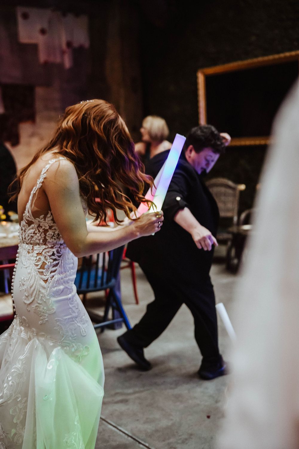 Bride in lace mermaid gown dances with glowing foam stick at evening wedding reception celebration.