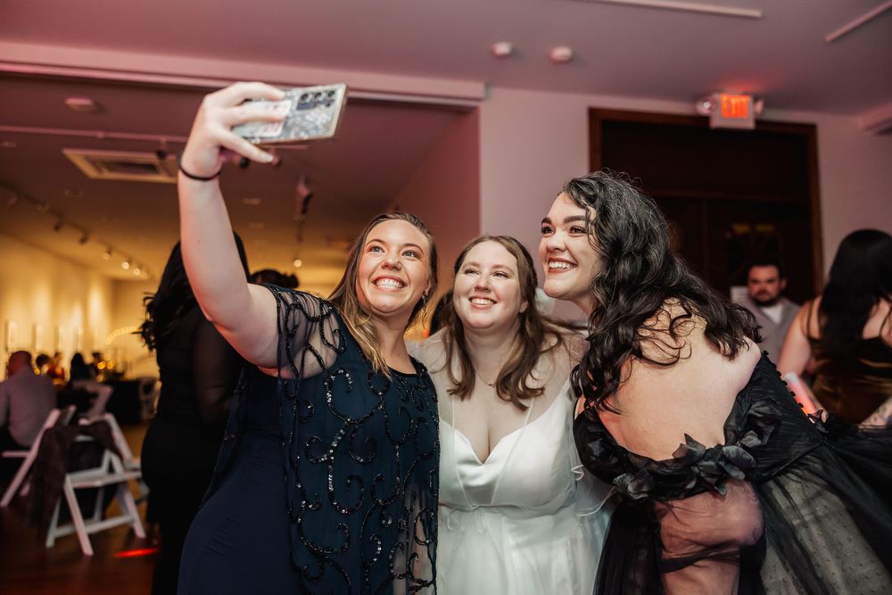 Three women smile and take a selfie together at a formal wedding reception event.
