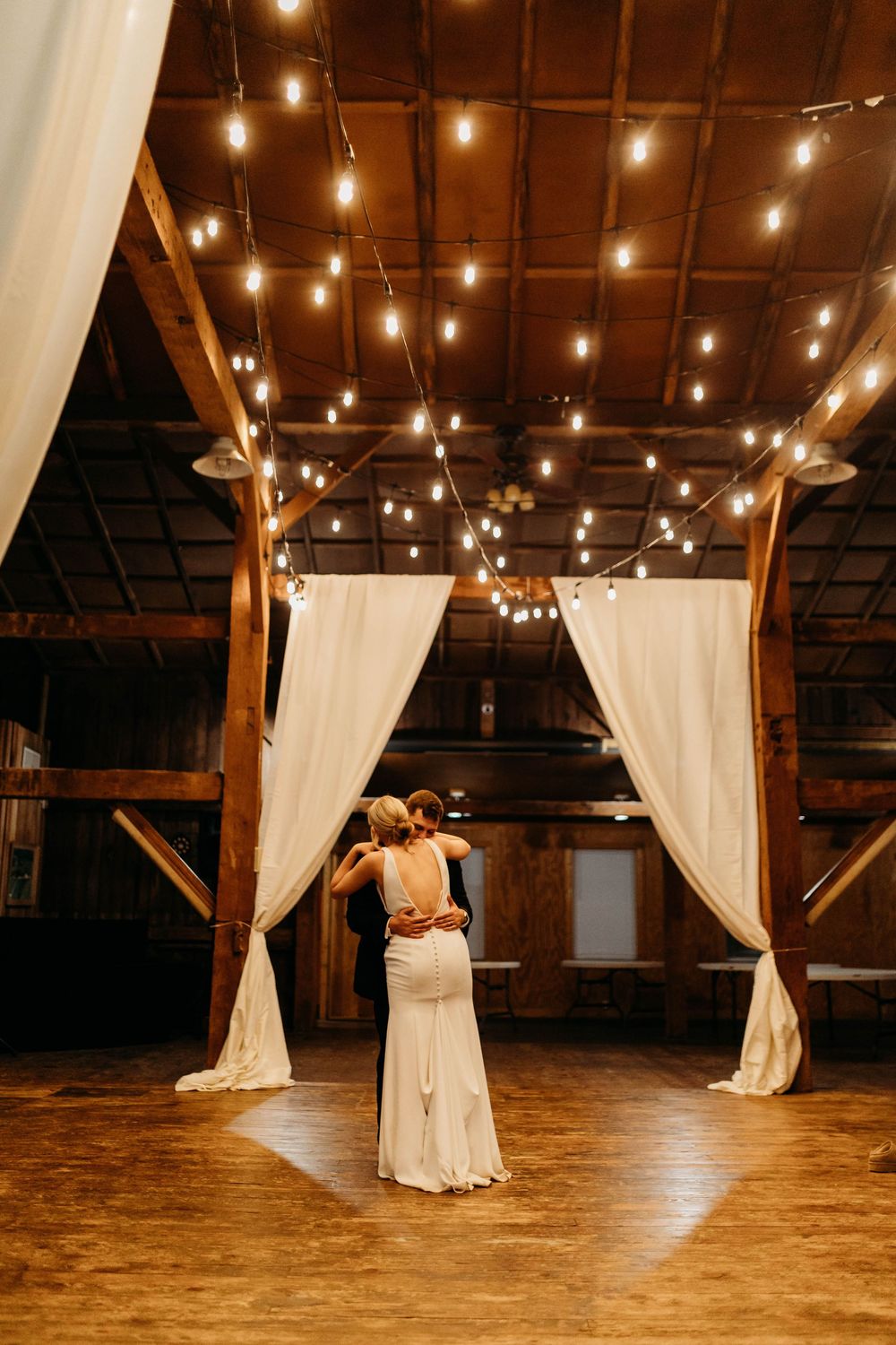 Couple sharing first dance in rustic barn wedding venue with string lights and white draping curtains.