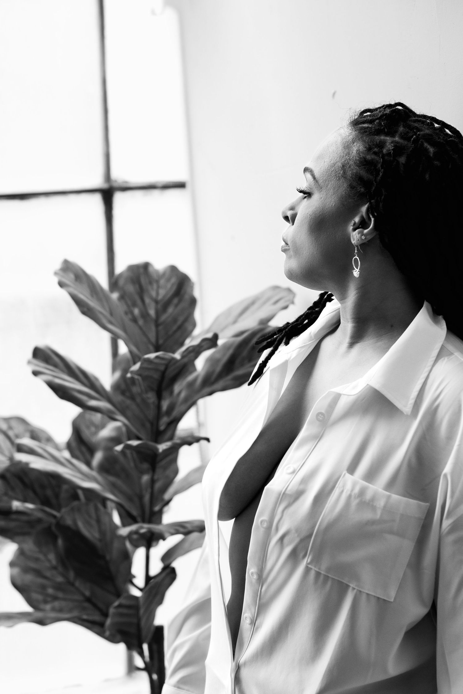 Black and white portrait of a woman in an open white shirt beside a fiddle leaf fig plant, looking up gracefully.