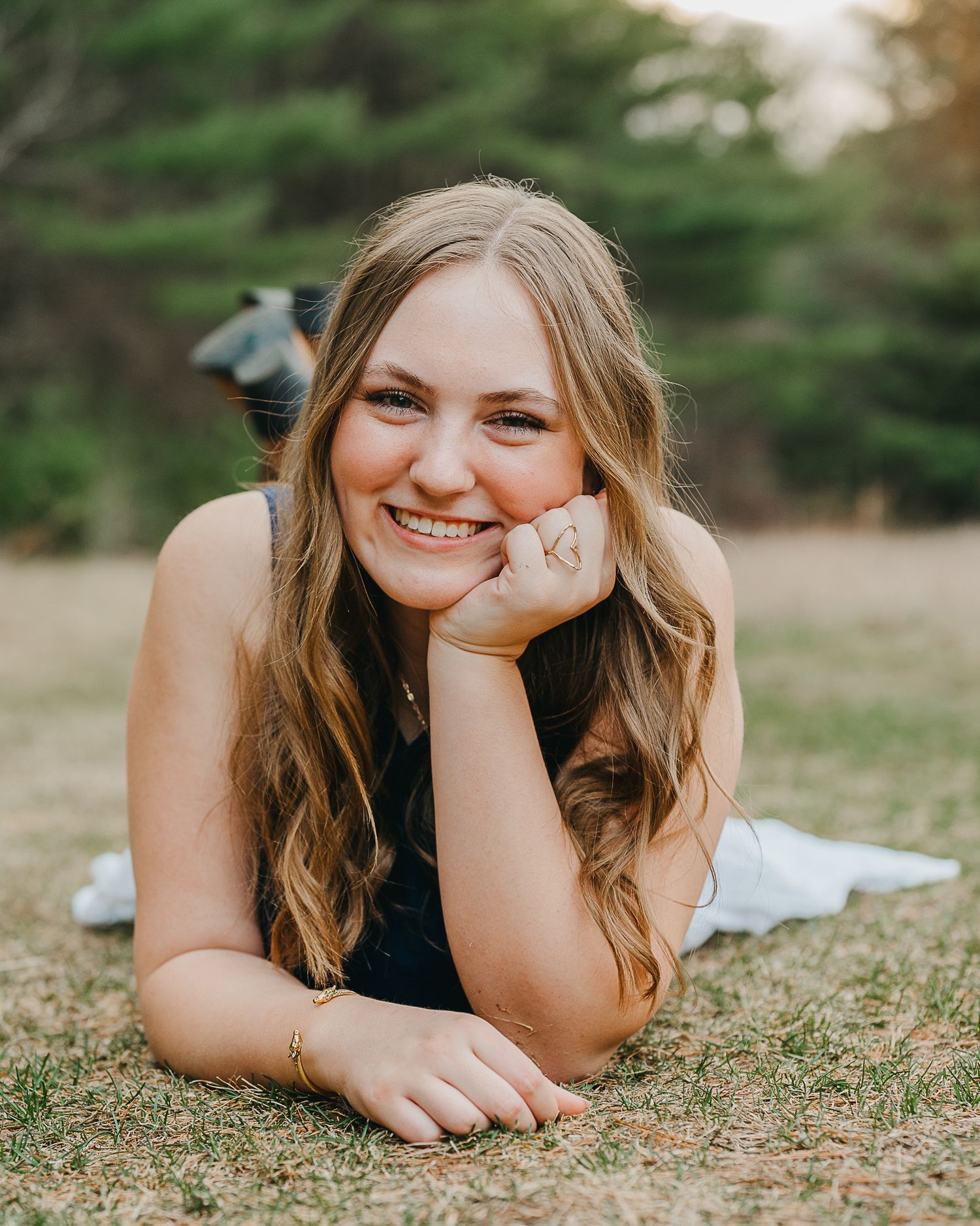 Smiling young woman with long wavy hair lying on grass in a park, resting chin on hand, wearing a black top.