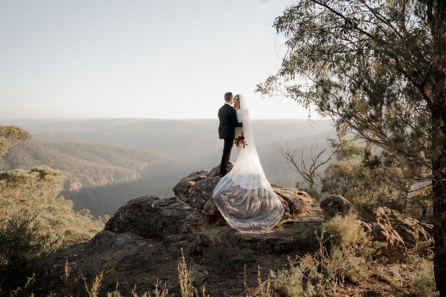 Bride and groom stand on rocky cliff edge with flowing veil, overlooking vast mountain valley landscape.