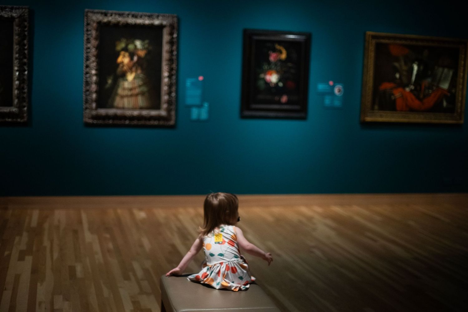 Young girl sitting on a bench in an art museum, admiring colorful paintings on a teal wall.