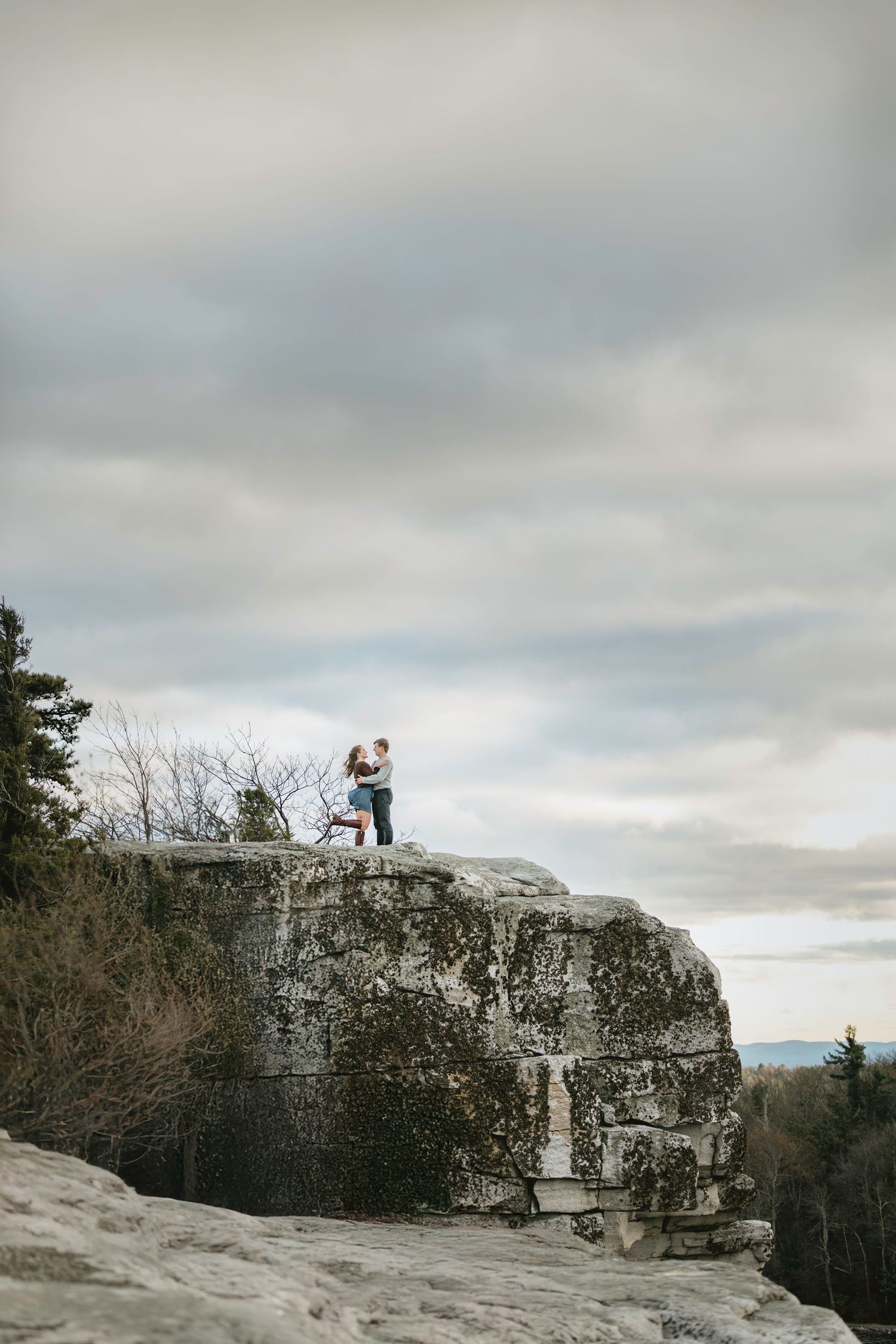 Couple embracing on a rocky cliff edge surrounded by trees under a dramatic cloudy sky.