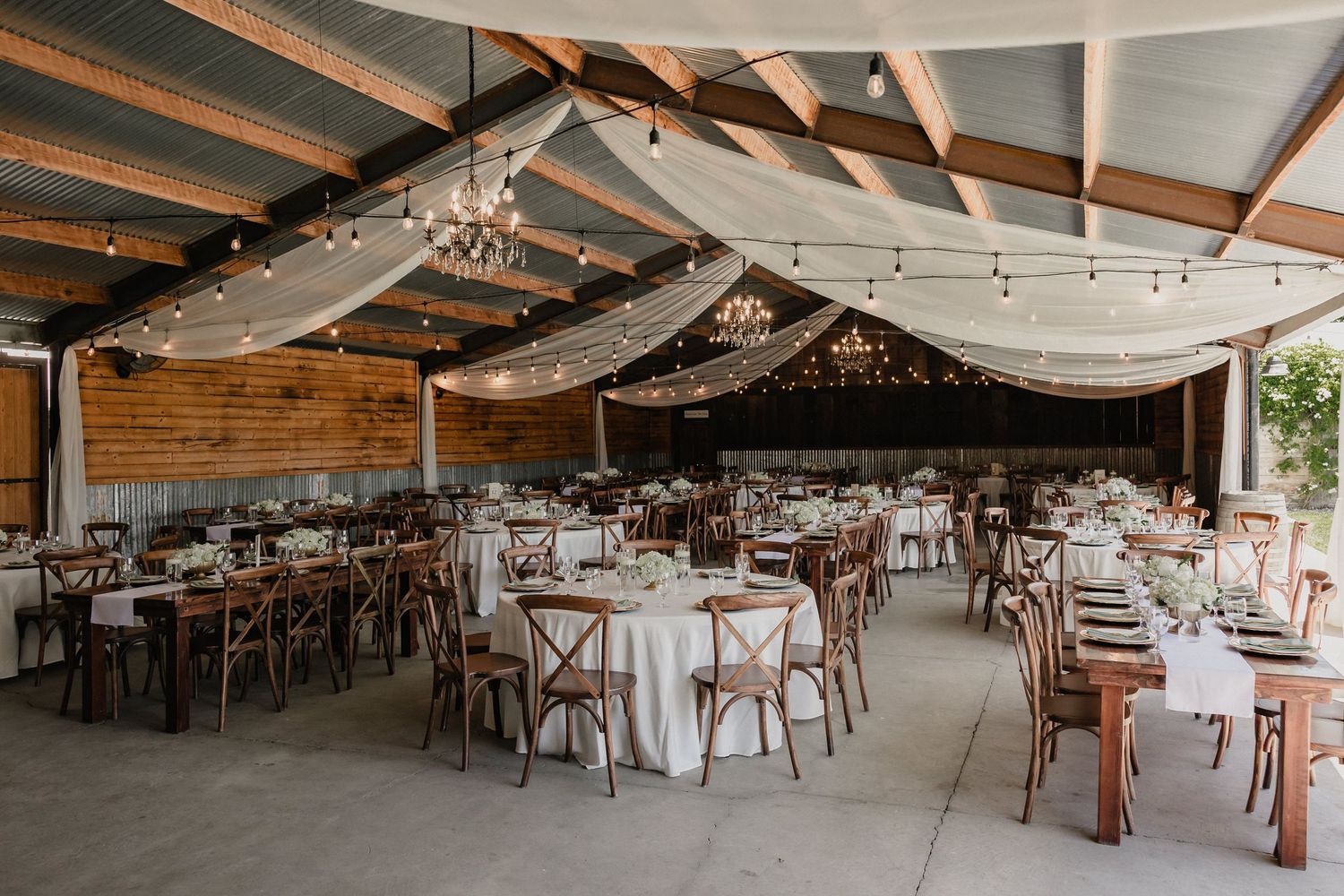 Elegant barn wedding reception with draped ceilings, string lights, chandeliers, and rustic wooden tables.
