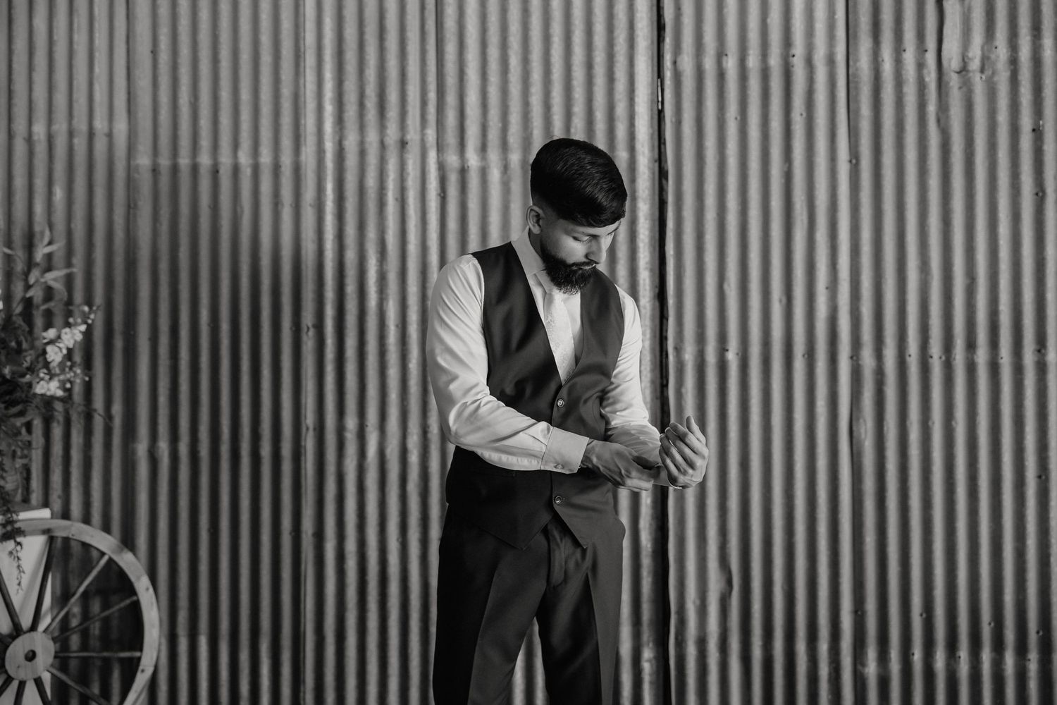 Groom in black vest adjusts cufflinks against corrugated metal wall in black and white photo.