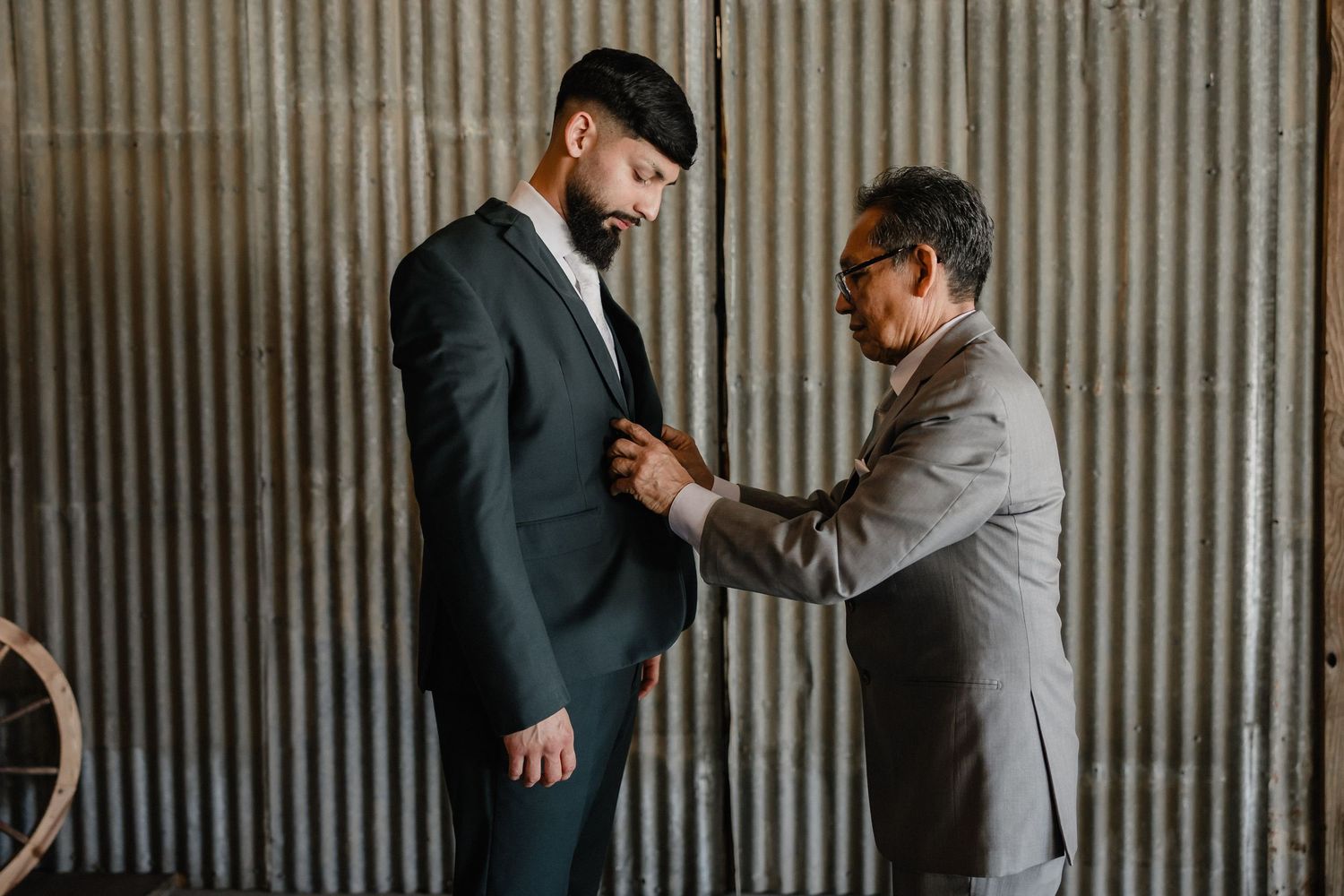 An older man in a gray suit adjusts the jacket of a younger groom in a dark suit before a wedding.