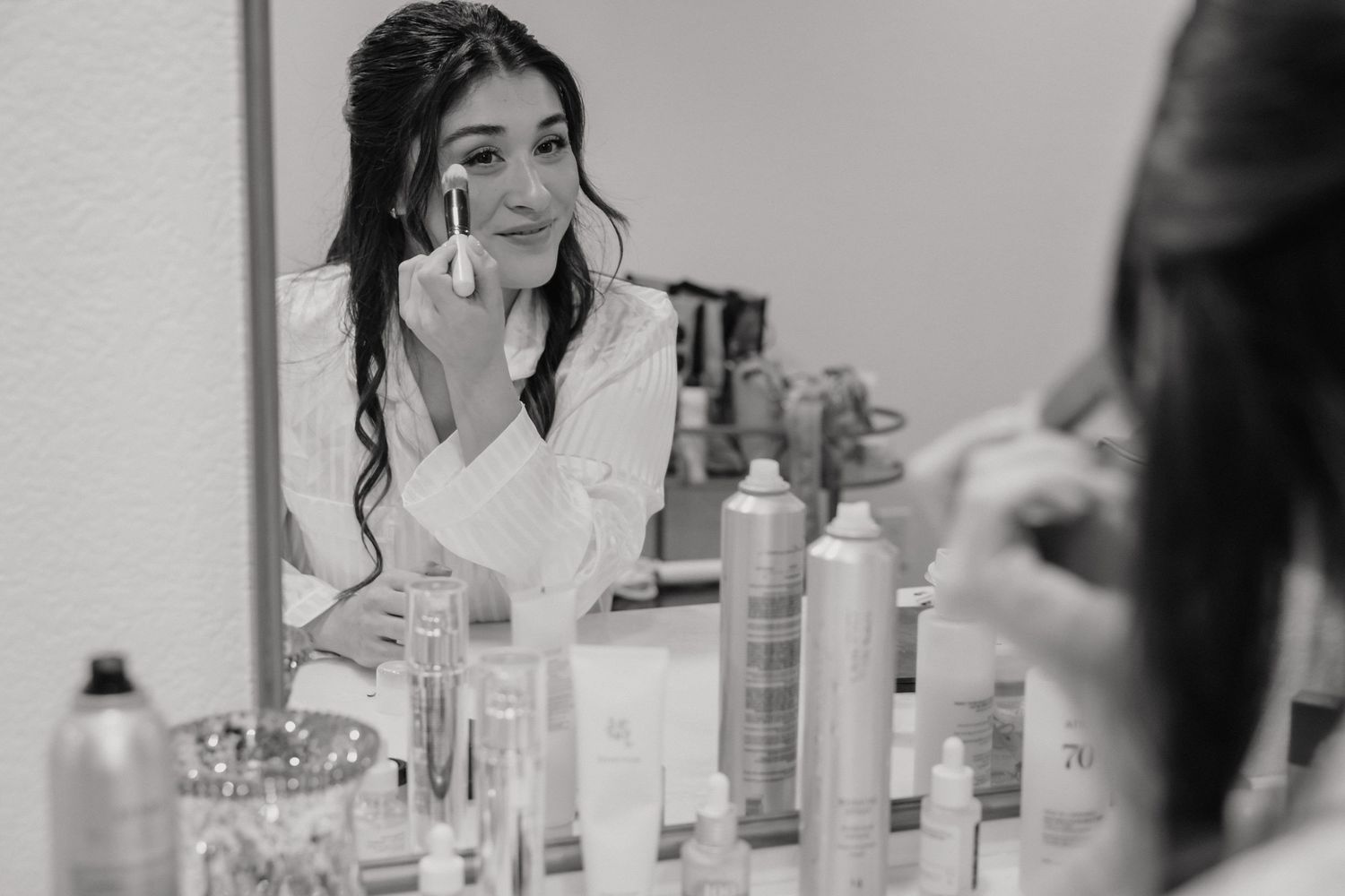 Black and white photo of bride applying makeup while looking in mirror during wedding preparations.