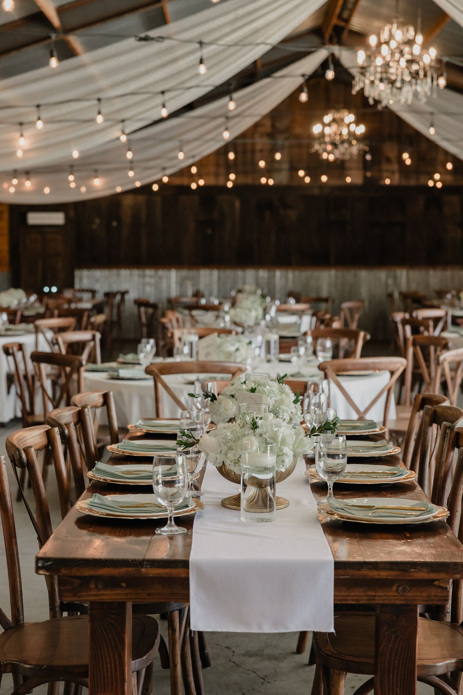 Rustic barn wedding reception with string lights, chandelier, wooden tables, white floral centerpieces, and elegant place settings.